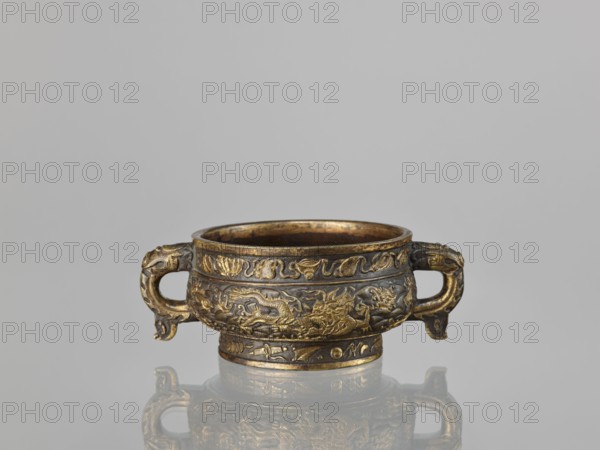 Gui-type incense burner hu. Maker: Hu, Wenming fl. ca. 1583–1614. Chinese; 19th century, Qing dynasty. Porcelain/ceramic ritual incense vessel decorated with dragons, phoenixes, unicorns qilin, plant ornament, hundred antiquities baigu motif, and sea waves. Height 7 cm; width 18.6 cm; diameter 13.1 cm., A small round metal vessel with two horizontally oriented handles is shown against a neutral gray background, resting on a reflective surface that mirrors its underside and handles; the vessel is richly decorated with raised ornamental patterns and relief figures, including swirling motifs and stylized animal or dragon-like forms, covering its body and base; the metal shows a mix of warm golden highlights and darker aged or patinated areas, giving an overall appearance of gold and dark brown tones, with subtle variations where the raised decoration catches the light.