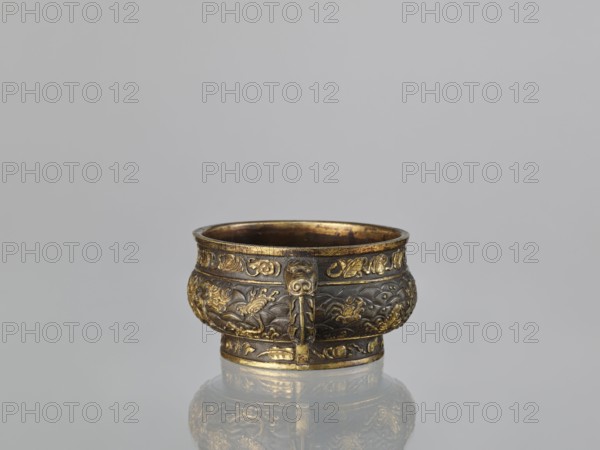 Gui-type incense burner hu, attributed to Hu Wenming fl. ca. 1583–1614. Chinese, 19th century, Qing dynasty. Bronze ritual vessel used as an incense burner. Decorated with dragons, phoenixes, unicorns qilin, plant ornament, sea waves, and hundred antiquities baigu motifs. Height 7 cm; width 18.6 cm; diameter 13.1 cm., An ornate metal bowl sits centered against a plain light gray background, its surface showing a mix of dark bronze and bright gold tones; the bowl has a rounded body with a slightly flared rim and a short base, and it features raised decorative bands and embossed motifs around its sides, including flowing wave-like patterns and stylized organic shapes; a vertical decorative strip with a face-like element projects slightly from the front, and the interior and rim show a warm golden sheen; the bowl rests on a reflective surface that mirrors its colors and shapes.