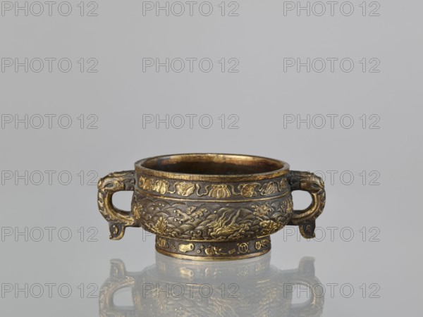 Gui-type incense burner hu, attributed to Hu Wenming fl. ca. 1583–1614. Chinese; Qing dynasty, 19th century. Bronze ritual vessel for incense, height 7 cm, width 18.6 cm, diameter 13.1 cm. Decorated with dragons, phoenixes, unicorns qilin, plant ornament, sea waves, and hundred antiquities baigu motif., An ornate, low round vessel with two short handles sits centered on a reflective surface, showing a mirrored image below; the vessel is decorated with raised, intricate motifs and relief patterns, including stylized animal and organic forms, and has a banded rim and foot with further decorative detailing. The visible colors are gold and dark brown on the vessel, with reflections of those tones on the surface, set against a light gray background.