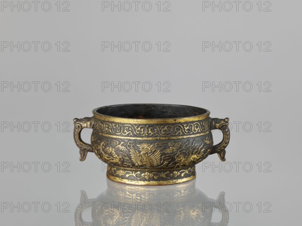 Gui-type incense burner, Chinese, Qing dynasty 1644–1911, 19th century. Bronze vessel with plant tendril ornamentation, decorated with phoenixes, dragons, unicorns qilin, and cranes. Ceremonial/ritual vessel. Height 7.5 cm; width 17.8 cm; diameter 13.7 cm; weight 0.53 kg., A small metallic bowl with a pair of ornate handles sits on a reflective surface, showing a faint mirrored image beneath it. The bowl is primarily dark metal with extensive gold-toned decorative accents and inlaid patterns around its rim, body, and base. The body features raised decorative motifs including stylized bird- or dragon-like figures and flowing ornamental designs. The handles are curved and sculpted with decorative detailing. The interior of the bowl appears dark and worn. The background is a uniform light gray. Colors visible include dark metal tones, gold, and light gray.