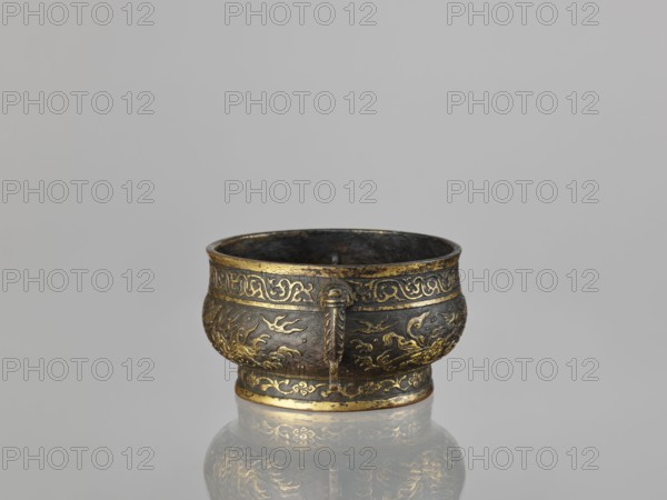 Gui-type incense burner, Chinese, Qing dynasty 1644–1911, 19th century. Bronze vessel with phoenixes, dragons, qilin unicorns, cranes, and plant tendril ornament; ceremonial/ritual function. Dimensions: height 7.5 cm; width 17.8 cm; diameter 13.7 cm; weight 0.53 kg., An ornate metal bowl sits on a reflective surface against a plain light gray background, the bowl showing a dark bronze base color with decorative gold accents and patterns around its rim and body, a slightly flared lip, a rounded body with embossed motifs and a small vertical decorative tab on one side, and the reflection of the bowl visible on the surface beneath it.