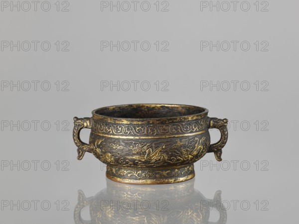 Gui-type incense burner, Chinese, Qing dynasty 1644–1911, 19th century. Bronze vessel with phoenixes, dragons, unicorns qilin, cranes, plant tendrils and other plant ornament; ceremonial/ritual function. Dimensions: height 7.5 cm; width 17.8 cm; diameter 13.7 cm; weight 0.53 kg., A small ornate metal bowl with a pair of handles sits centered against a plain light gray background, showing a reflective surface beneath it. The bowl is dark with areas of golden highlights and worn bronze tones, decorated with raised swirling patterns and stylized animal motifs encircling its body. The handles curve outward and have carved details with the same dark and gold coloration. The rim and base show gilded accents and some darker patina, and the bowl’s interior appears dark with subtle gold flecks. The reflection below repeats the bowl’s dark, gold, and bronze colors on the glossy surface.