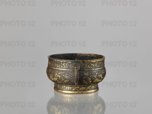 Gui-type incense burner, Chinese, Qing dynasty 1644–1911, 19th century. Bronze ceremonial/ritual vessel with phoenixes, dragons, cranes, unicorns qilin, plant tendril ornamentation. Height 7.5 cm; width 17.8 cm; diameter 13.7 cm; weight 0.53 kg., A small metal bowl sits centered on a smooth reflective surface, showing a mirrored reflection beneath it; the bowl is primarily dark bronze with ornate gilded accents in gold, featuring embossed decorative patterns and raised motifs around its body and rim, and it has a short rounded foot with similar gold detailing; the interior appears darker and aged, while the background is a plain light gray.