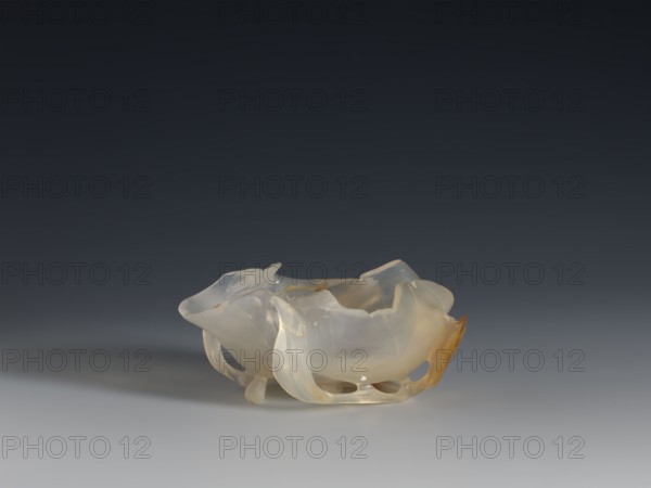 A vessel for washing brushes, Chinese, Qing dynasty 1644–1911, 18th century. Sculpture: a bowl resembling a leaf, entwined on the outside with a branch with small leaves; plant ornament; used with writing implements. Vessel, height 3.8 cm, width 8.3 cm., A translucent, irregularly shaped bowl-like object with smooth, curved edges and carved openings sits on a flat surface; its surface shows a mix of pale colors including milky white, cream, and light amber with subtle variations and glossy reflections, against a gradient background that transitions from dark gray at the top to lighter gray near the surface.