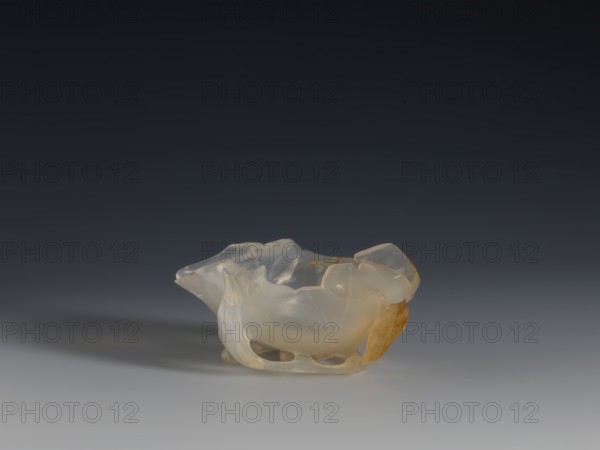 A vessel for washing brushes, Chinese, Qing dynasty 1644–1911, 18th century. Sculpture: a bowl resembling a leaf, entwined on the outside with a branch with small leaves; ornament of plants; related to writing implements. Vessel bowl, height 3.8 cm, width 8.3 cm., A translucent sculpted object sits on a smooth surface against a dark gradient background, appearing in pale cream and milky white tones with patches of warm amber and light brown. The object has an organic, flowing shape with curved, undulating edges and several hollowed openings along its base, creating soft highlights and subtle internal reflections. The surface shows smooth, glossy areas and gentle translucency that reveal layered color variations, and a soft shadow extends from the object onto the surface.