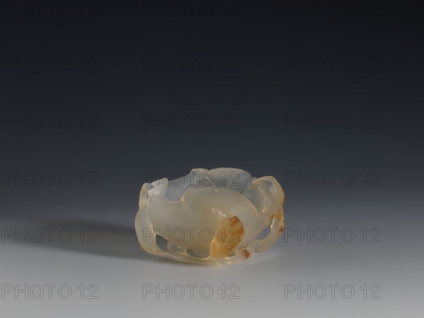 A vessel for washing brushes, Chinese, Qing dynasty 1644–1911, 18th century. Vessel — a bowl resembling a leaf, entwined on the outside with a branch with small leaves; used for washing writing implements. Sculpture, vessel; height 3.8 cm, width 8.3 cm., A small translucent bowl-like object with an irregular scalloped rim sits on a smooth surface, showing a mix of pale milky white and warm amber tones with areas of clearer, nearly colorless translucency; the object has carved openings and curved petal-like forms, and casts a soft shadow to one side against a gradient background that transitions from dark gray at the top to lighter gray near the bottom.