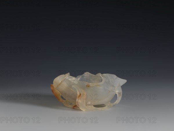 A vessel for washing brushes, Chinese, Qing dynasty 1644–1911, 18th century. Vessel — a bowl resembling a leaf, entwined on the outside with a branch with small leaves; ornament: plant motif; function: writing implements vessel. Sculpture, height 3.8 cm, width 8.3 cm., A translucent sculptural object rests on a smooth surface against a dark gradient background; the object has an organic, rounded form with flowing, curved edges and several cutout openings, appearing smooth and slightly glossy. The colors visible are pale milky white, warm amber tones, and hints of soft beige, with subtle variations where the material is thinner or thicker. Shadows fall beneath and slightly to the left of the object, and the background transitions from very dark near the top to a muted gray near the surface.