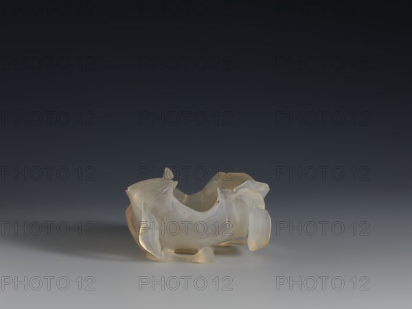 A vessel for washing brushes, 18th century, China, Chinese culture, Qing 1644–1911, Qing style. Sculpture; vessel – a bowl resembling a leaf, entwined on the outside with a branch with small leaves; ornament: plant; related to writing implements. Height 3.8 cm; width 8.3 cm., A translucent carved object with smooth, curving forms and irregular edges sits on a flat surface, showing shades of pale white, creamy beige, and faint amber, with soft reflections and a subtle shadow extending from it against a gradient background that transitions from dark gray at the top to lighter gray near the surface.
