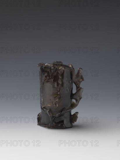 Vase on base, carved ornaments of flowers, birds, and stylized rocks; wooden oval base on three legs. Chinese, Qing dynasty 1644–1911, 18th–19th century. Vessel, height 11.8 cm, diameter 6.3 cm. Maker, Timoshka aka Timoshev, Nicholas., A dark translucent sculpted object sits against a gradient gray background, showing a glossy surface with smooth and carved areas; the piece has organic, flowing forms that suggest twisted branches and leaf-like shapes, and a bird-like figure is perched on one side. The colors visible are dark brown to almost black in the object, with highlights reflecting lighter brown and gray, and the background ranges from dark charcoal gray at the top to pale gray near the bottom.
