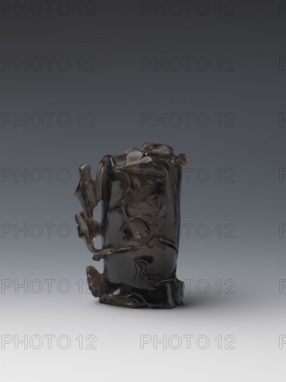 Vase on base, carved ornaments, flowers, birds decorative motifs, stylized rocks; wooden base, oval on three legs; Qing style Qing dynasty, 1644–1911; vessel; 18th–19th century China; height 11.8 cm, diameter 6.3 cm., A dark, glossy carved object resembling a short vertical cylinder with irregular contours sits on a smooth light gray surface against a gradient gray background; the object has organic carved motifs with flowing curved lines and raised rounded forms, and features protruding elements on one side that create openings and negative space, with visible color variations in deep brown to near-black and small areas of lighter brown translucence.