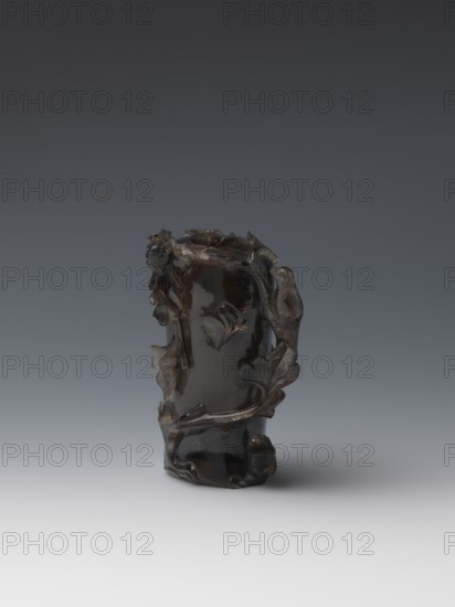 Vase on base, carved ornaments; wooden base, oval on three legs. Carved decoration: stylized rocks, flowers, birds. Chinese art; Qing style Qing dynasty, 1644–1911. 18th–19th century, China. Vessel. Height 11.8 cm; diameter 6.3 cm., The image shows a carved dark brown translucent object with an irregular organic shape, featuring sculpted leaf-like and vine-like forms wrapping around its sides and a small rounded protrusion near the base; the object sits on a smooth surface and is lit against a gradient background that transitions from dark gray at the top to light gray at the bottom, with visible colors including dark brown, gray, black, and hints of lighter brown in the object's surface reflections.