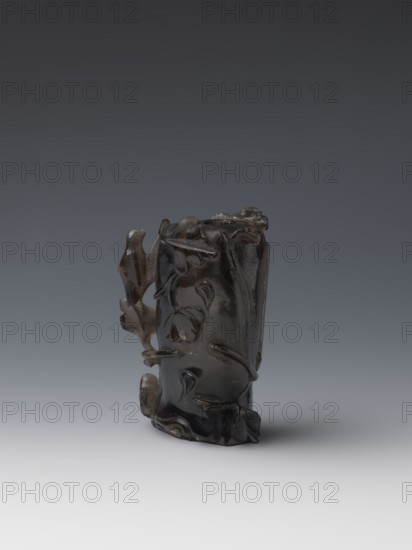 Vase on base, carved ornaments, carved decoration: stylized rocks; flowers ornament; birds decorative motif; Qing style, Qing 1644–1911; 18th–19th century; China; wooden base, oval on three legs; vessel; height 11.8 cm, diameter 6.3 cm;, A dark, glossy carved object with an irregular, organic shape sits centered on a smooth surface; the carving shows flowing, rounded forms and leaf-like details with protruding elements and textured surfaces, and the overall colors visible are dark brown to black with subtle lighter brown highlights against a gradient background that transitions from dark gray at the top to light gray at the bottom.