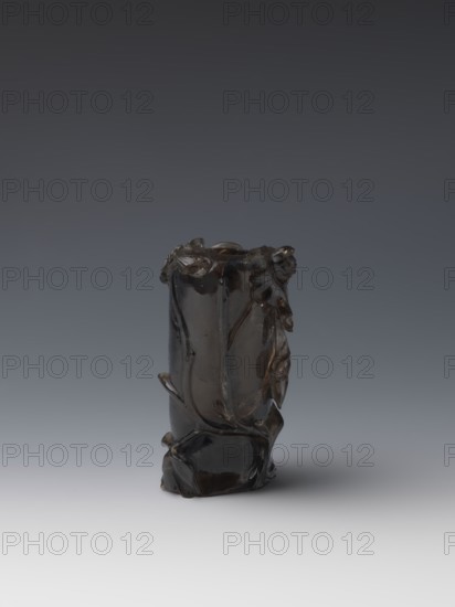 Vase on base, carved ornaments, flowers and birds decorative motifs, stylized rock carving; wooden oval base on three legs. Chinese, Qing dynasty 1644–1911, 18th–19th century. Vessel, height 11.8 cm, diameter 6.3 cm., A dark brown glossy carved object with irregular, organic contours standing upright on a smooth surface, featuring flowing, rounded protrusions and carved leaf-like forms that wrap around its cylindrical body, with highlights reflecting off its polished surface and subtle variations in brown and almost black tones against a neutral gradient gray background.