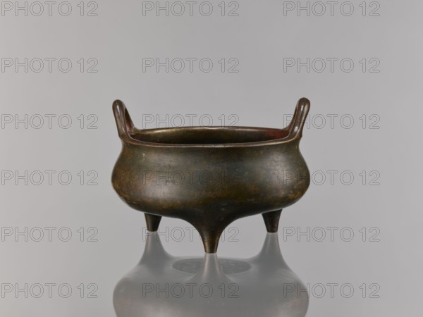 Li-type incense burner, bronze, Chinese, Qing dynasty 1644–1911, 18th–19th century, ceremonial/ritual vessel with dragon iconography, incense burner, height 13.5 cm, width 18.6 cm., A low, round metal vessel with a dark brown and greenish patina stands on three tapered legs, with a pair of short upward-curving handles at the rim; the interior shows hints of reddish tones and the vessel casts a soft reflection on a glossy surface beneath it, set against a pale gray background.