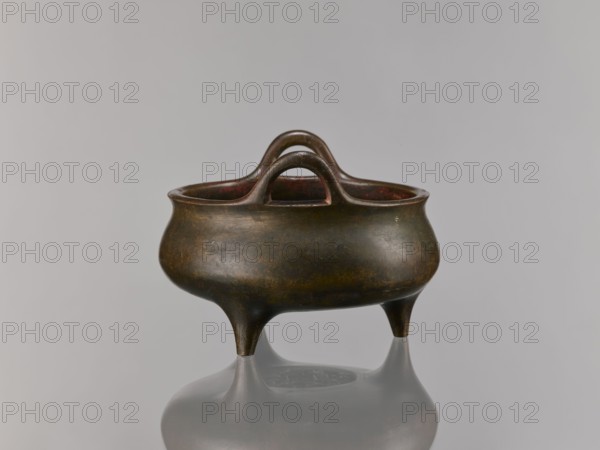 Li-type incense burner, bronze, 18th–19th century Qing dynasty 1644–1911, China. Ceremonial/ritual vessel with dragon iconography; height 13.5 cm, width 18.6 cm., A small rounded vessel with a patinated bronze surface in shades of dark brown, greenish-brown, and hints of reddish-brown sits on three short tapered legs, showing a smooth curved body and a slightly flared rim; a pair of arching handles rise from opposite sides and meet above the center, and the vessel casts a soft reflection on a pale gray reflective surface against a light gray background.