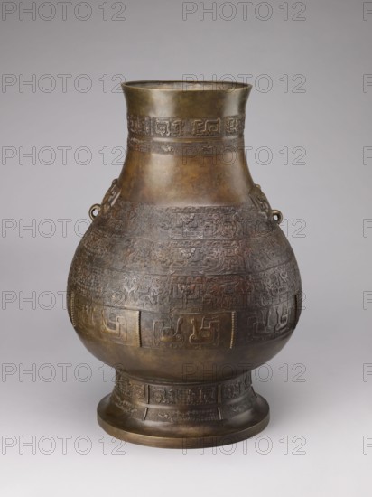 Hu-type vase, Chinese art, Chinese culture; birds; ceremonial/ritual vessel; dragons iconography; taotie iconography; Chinese symbols; Qing dynasty 1644–1911. A hu-type ritual vessel with an ovoid-shaped belly, a cylindrical neck straight at the rim, and a high, distinct foot with a conical profile. Semicircular clubs are attached to the collar. Rich relief decoration on the belly emphasized with engraving. Classic decorative motifs: taotie masks, mythical kui animals, stylized birds, and geometric patterns. 18th–19th century, China; vessel ritual. Height 59.5 cm; width 40.0 cm; weight 19.0 kg., An aged metal vessel with a rounded body, a flared neck, and a footed base sits against a neutral background; the surface shows intricate low-relief patterns and decorative bands circling the body and neck, and small ring handles project from the shoulder area. The visible colors include brown, dark bronze, muted gold, and hints of greenish patina, with subtle variations of darker and lighter b...