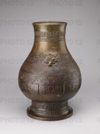 Hu-type vase, Chinese, Qing 1644–1911, 18th–19th century. A hu-type ritual bronze vessel with an ovoid-shaped belly, cylindrical neck with a straight rim, and a high conical foot. Semicircular clubs are attached to the collar. Rich relief decoration on the belly emphasized with engraving; classic decorative motifs include taotie masks, mythical kui animals, stylized birds, dragons, Chinese symbols, and geometric patterns. Ritual vessel, height 59.5 cm, width 40.0 cm, weight 19.0 kg., A large rounded metal vessel with a flared neck and a pedestal base sits against a neutral gray background; the vessel displays a dark bronze and brown patina with areas of lighter gold-toned metal, and its surface is covered in intricately carved or cast decorative bands and raised motifs including a stylized animal or mask-like form on the upper body, with repeating geometric and curvilinear patterns encircling the midsection and base, and subtle variations of darker brown, reddish-brown, greenish-brown,...