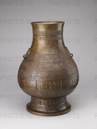 Hu-type vase, Chinese, Qing 1644–1911, 18th–19th century. A hu-type ritual bronze vessel with an ovoid-shaped belly, a cylindrical neck straight at the rim, and a high conical-foot. Semicircular clubs attached to the collar. Rich relief decoration on the belly emphasized with engraving. Classic decorative motifs: taotie masks, mythical kui animals, stylized birds, dragons, Chinese symbols and geometric patterns. Ritual vessel; height 59.5 cm, width 40.0 cm, weight 19.0 kg., A large rounded metal vessel with a narrow flared neck and a wide foot, shown against a plain light gray background. The vessel has an overall aged brown and bronze patina with darker and lighter areas, and a few greenish and reddish tints visible on the surface. The body is decorated with several horizontal bands of low-relief geometric and stylized motifs, and there are a few raised loop handles on the shoulder. The foot and neck also feature banded decorative patterns. The surface shows subtle unevenness, small d...