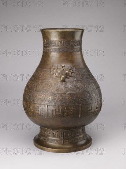 Hu-type vase, Chinese art, Chinese culture; subject: birds, dragons, taotie, Chinese symbols; function: ceremonial/ritual vessel bronze hu-type; description: ovoid-shaped belly, cylindrical neck with straight rim, high distinct conical foot; semicircular clubs attached to the collar; rich relief decoration on the belly emphasized with engraving; classic decorative motifs: taotie masks, mythical kui animals, stylized birds, geometric patterns; period: Qing 1644–1911, 18th–19th century, China; dimensions: height 59.5 cm, width 40.0 cm, weight 19.0 kg., A large, rounded metal vessel with a narrow flared neck and a broad foot stands against a plain light gray background; the vessel shows an overall brown and bronze patina with areas of darker and lighter bronze tones, and a few subtle greenish and golden highlights. The body of the vessel is covered in repeated raised decorative bands and intricate carved or cast geometric and zoomorphic patterns encircling the midsection and neck, and a s...