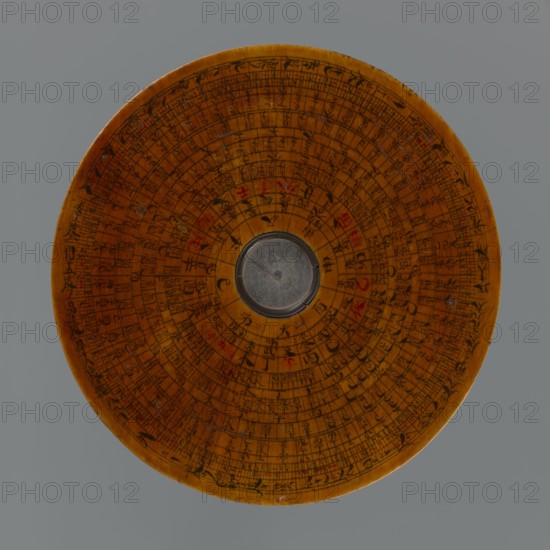 Feng shui compass luopan, navigational object; Chinese; Qing dynasty 1644–1911, 19th century; Huizhou, China; measuring instrument; diameter 13.8 cm; includes calendars, clocks, and Chinese Zodiac markings., A round wooden disc with concentric engraved rings and a small circular metal piece set at the center, the wood showing warm brown and amber tones with darker brown engraved markings and occasional red accents, all set against a flat gray background.