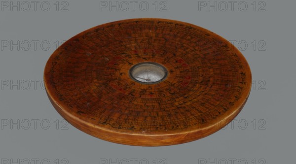 Feng shui compass luopan, navigational object; Chinese; Qing dynasty 1644–1911, 19th century; Huizhou, China; materials: measuring instrument; diameter 13.8 cm; includes calendars, clocks, and Chinese Zodiac signs., A round wooden disc with a warm brown surface and a slightly darker rounded edge sits against a neutral gray background; the top face shows concentric circular patterns and numerous fine dark markings and short strokes arranged in rings, with small areas of red accents scattered across the surface; a shallow circular metal-rimmed recess with a reflective silver interior is set near the center, and the wood displays subtle variations in grain and glossy wear.