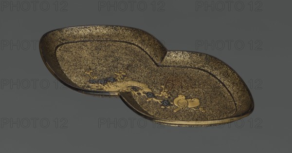 Tray, lacquerware, Japanese art; subject: plum trees and birds kachôga; decoration: asymmetrical composition of an old plum tree trunk with young shoots; function: vessel/tray; period: 1st half of the 19th century Edo period, 1603–1868; origin: Japan; dimensions: height 2.6 cm, width 17.8 cm, depth 9.5 cm., A shallow, decorative dish with a double-lobed shape sits against a dark gray background; the dish surface is predominantly gold with a textured, speckled appearance and darker brown or black edges and outlines, and it features a raised ornamental design including a stylized plant motif and a pair of small birds in a slightly brighter gold tone with some darker accents.