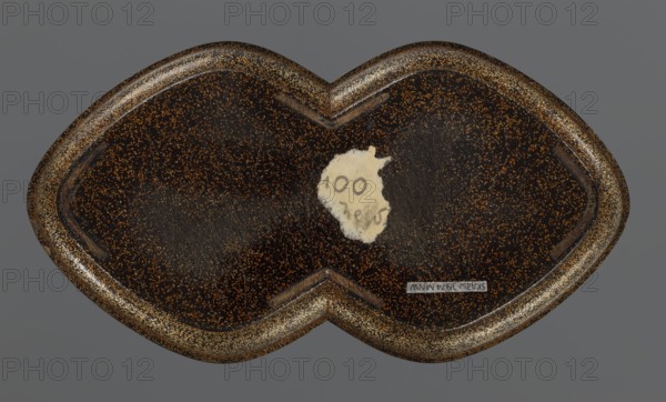 Tray, lacquerware, Japanese art; tray with asymmetrical decoration of an old plum tree trunk with young shoots and birds kachôga decorative motif; Edo period, 1st half of the 19th century; Japan; vessel; height 2.6 cm, width 17.8 cm, depth 9.5 cm., A close-up view of a heart-shaped object with a dark brown surface speckled with gold or tan flecks, set against a flat gray background; the object has a glossy finish with visible surface scratches and a worn spot revealing a lighter beige area, and its raised rim shows denser gold flecking and a slightly darker brown edge.