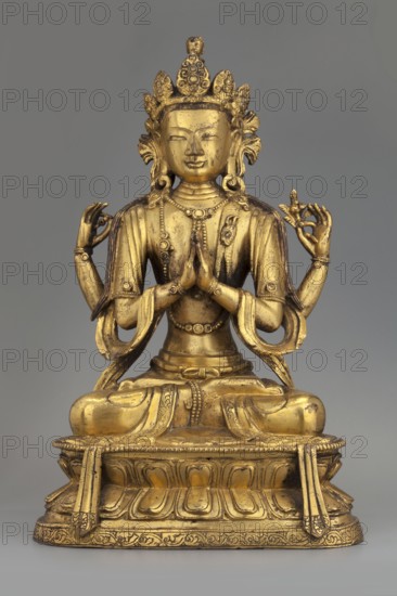 Four-armed Bodhisattva Avalokiteshvara Chaturbhuja, The Bodhisattva shown in one of his tantric forms called shadakshari. A single-headed, four-armed figure, seated in a meditative pose on a lotus throne. One pair of hands is in the namaskara mudra; the other holds attributes: a lotus flower and a rosary almost unpreserved, only one element remains. Dressed in typical bodhisattva regalia: a crown, earrings, necklace, inlaid bracelets on wrists and ankles, a sash flowing from shoulders to forearms, an antelope skin draped over the left shoulder, and a foot-long bandage with a floral pattern around the lower body. A coiffed knot of hair atop his head is crowned by the head of Amitabha. The plaque closing the base is missing. 2nd half of the 18th century. China/Tibet. Sculpture. Height 22.5 cm, width 14.5 cm, depth 9.5 cm., A gilded statue of a seated figure with a crown and multiple arms is shown against a neutral gray background; the figure sits on an ornate pedestal with carved floral ...