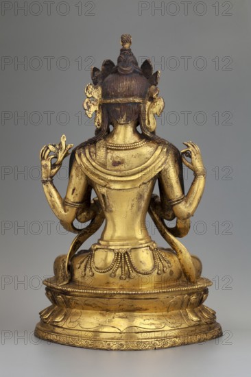 Four-armed Bodhisattva Avalokiteshvara Chaturbhuja, Buddhist/Tibetan/Himalayan/Sino-Tibetan/Chinese art. The Bodhisattva shown in a tantric form called shadakshari: single-headed, four-armed figure seated in meditation on a lotus throne. One pair of hands in the namaskara mudra; the other holds a lotus flower and a rosary almost unpreserved apart from one element. Dressed as a bodhisattva with a crown, earrings, necklace, inlaid bracelets on wrists and ankles, a sash flowing from shoulders to forearms, an antelope skin draped over the left shoulder, and a foot-long bandage with a floral pattern around the lower body. A coiffed hair knot is topped by the head of Amitabha. Plaque closing the base is missing. 2nd half of the 18th century. China/Tibet. Sculpture. Height 22.5 cm; width 14.5 cm; depth 9.5 cm., The image shows the rear view of a gold-colored statue seated on an ornamental base against a plain gray background, with a few areas of darker patina; the figure has a dark brown hair...