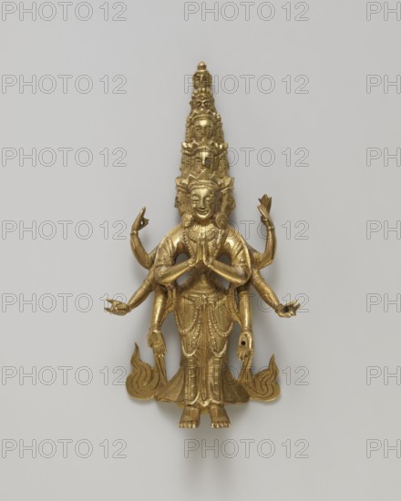 Guanyin, figurine, Chinese/Tibetan art, bodhisattva iconography, heads, flames, hands; 19th century; Tibet/China possible origin; sculpture; height 9.9 cm, width 5.2 cm, depth 2.0 cm., A gilded metal figurine of a standing multi-armed deity with a tall, tiered headdress composed of several smaller faces stacked above the main head, the main face displaying a calm expression and ornate ear discs, a pair of hands held together at chest level while additional arms extend outward in varied gestures, the figure wearing a detailed draped garment with decorative patterns and beaded necklaces, the feet visible at the base standing on a stylized flame-like platform, the overall color palette dominated by gold against a neutral light gray background.