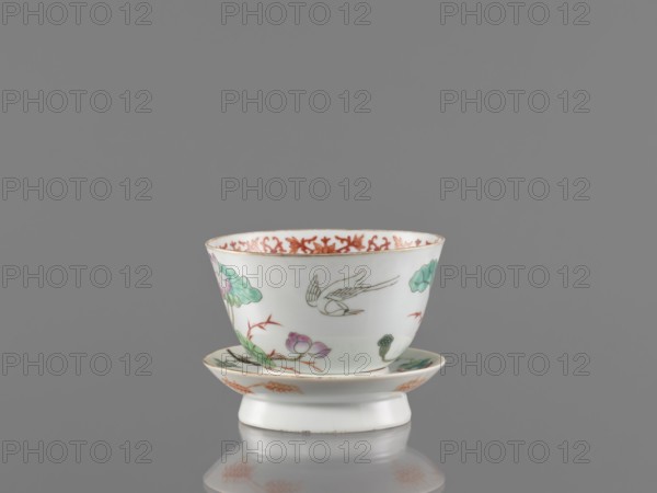 Bowl with stand, ceramics. Decorated colored porcelain in Guangxu style Qing dynasty, 19th/20th century, 1875–1908. Motifs: lotuses, cranes, bamboos; interior decorated with an ornamental band. Obverse of the stand shows a bird and flowered branches; reverse shows bamboo shoots. Chinese; vessel. Height 8.5 cm, diameter 10.6 cm., A white porcelain cup with a matching saucer set on a short pedestal, decorated with a painted bird in flight and floral motifs in green and pink, with accenting reddish-orange patterns along the rim and on the saucer, photographed against a plain gray background with a faint reflection beneath the pedestal.