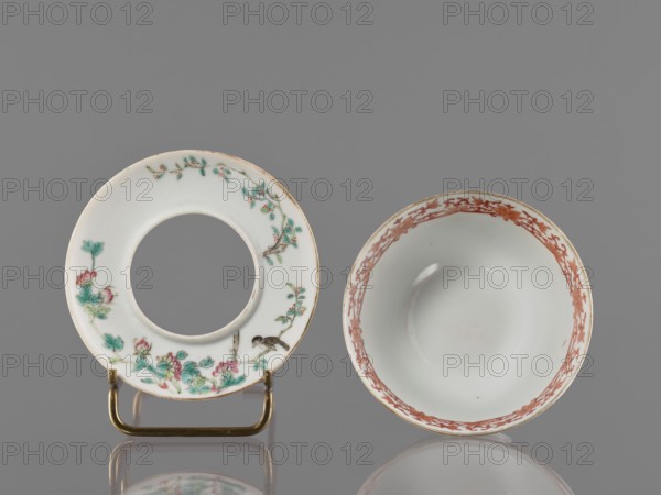 Bowl with stand, ceramics, colored porcelain, decorated inside with an ornamental band. On the obverse of the stand are a bird and flowered branches; on the reverse are bamboo shoots. Motifs: lotuses, cranes, bamboos. Style/period: Guangxu era, Qing dynasty 1644–1911, 19th/20th century 1875–1908. Chinese; vessel. Height 8.5 cm, diameter 10.6 cm., A circular white ceramic ring with painted green leaves, pink flowers, brown branches, and a small brown bird sits on a brass-colored stand next to a white ceramic bowl decorated with a reddish-brown floral border around its rim, all set against a gray background with a subtle reflection beneath them.
