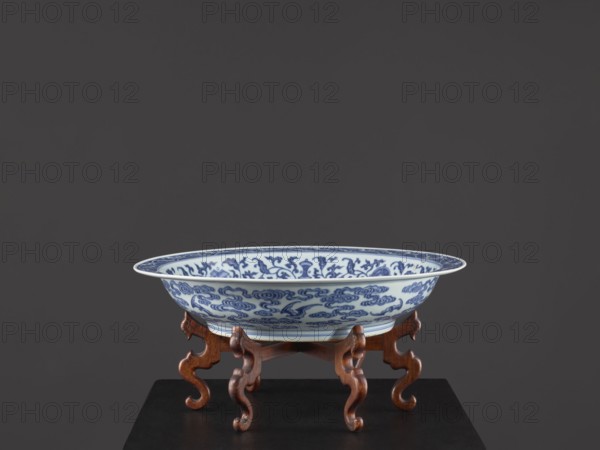 Platter on stand, Blue-and-white porcelain platter on a low foot with a wide rim, sitting on a carved wooden stand. Decorated with plant tendrils, bats, and Buddhist emblems. Center of the mirror features a rosette filled with an archaized swastika ornament; outer wall shows bats among clouds. Qing dynasty, Yongzheng period 1723–1735, China. Vessel; height 21 cm, diameter 45.5 cm., A shallow porcelain bowl with blue decorative patterns sits on a carved wooden stand; the bowl is primarily white with intricate blue designs on its interior and exterior, the stand is a warm brown wood with curving legs, and the background is a dark gray.