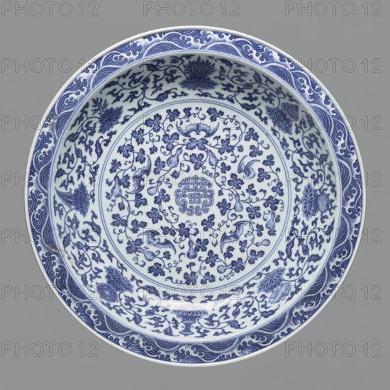 Platter on stand, Blue-and-white porcelain, Qing dynasty, Yongzheng period 1723–1735. Platter on a low foot with a wide rim, seated on a carved wooden stand. Decorated with plant tendrils, bats, and Buddhist emblems. Center of the mirror has a rosette filled with an archaistic swastika ornament; outer wall shows bats among clouds. China. Vessel; height 21 cm, diameter 45.5 cm., A round ceramic dish viewed from above with intricate blue floral and vine patterns on a white background, featuring a decorative central medallion surrounded by a ring of scrolling foliage and a border of stylized floral motifs; the rim has a wave-like patterned band in blue against white. The visible colors are blue, white, and gray.