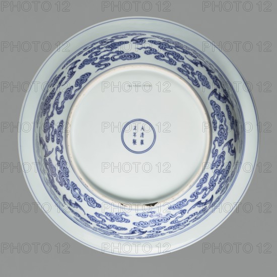 Platter on stand, decorated with bats and fruit motifs; blue-and-white porcelain, Qing dynasty, Yongzheng period 1723–1735. The platter on a low foot with a wide rim sits on a carved wooden stand. Decorated with plant tendrils, bats, and Buddhist emblems; the center mirror has a rosette filled with an archaized swastika ornament. Outer wall shows bats among clouds. China; vessel; height 21 cm, diameter 45.5 cm., A round porcelain bowl viewed from above with a glossy white interior and exterior, decorated with a repeated cloud-like pattern in deep blue along the inner sides and rim; the bowl has a pale blue band near the outer rim and a smooth central base with a small circular blue motif; visible colors include white, deep blue, pale blue, and a small area of dark brown on the rim.
