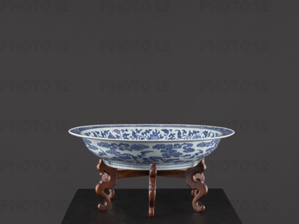 Platter on stand, Blue-and-white porcelain platter on a low foot with a wide rim, seated on a carved wooden stand. Decorated with plant tendrils, bats, and Buddhist emblems. Center of the mirror holds a rosette filled with an archaized swastika ornament; bats amid clouds on the outer wall. Qing dynasty, Yongzheng period 1723–1735. China. Vessel; height 21 cm, diameter 45.5 cm., A shallow round ceramic bowl with blue decorative patterns on a white background rests on a wooden stand with curved legs; the bowl shows ornate swirling motifs in blue, and the stand is a warm brown wood with carved, scroll-like legs; the surface beneath the stand is dark, and the background is a solid dark gray.