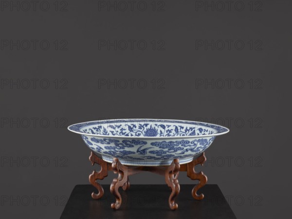 Platter on stand, blue-and-white porcelain, Qing dynasty, Yongzheng period 1723–1735. The platter on a low foot with a wide rim sits on a carved wooden stand. Decorated with plant tendrils with bats and Buddhist emblems. In the center of the mirror is a rosette filled with an archaized swastika ornament. On the outer wall there are bats among the clouds. Vessel, height 21 cm, diameter 45.5 cm. Chinese, ceramics, Yongzheng style., A shallow round ceramic bowl with blue decorative patterns on a white background sits on an ornate wooden stand; the bowl shows flowing floral and cloud-like motifs in blue, with a thin pale rim, and the stand is carved with curved legs in a warm brown wood tone, all set against a solid dark gray background and resting on a dark surface.