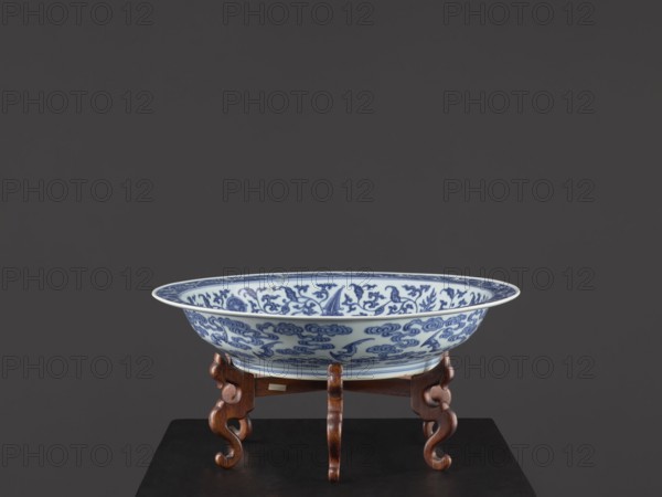 Platter on stand, Blue-and-white porcelain, Qing dynasty, Yongzheng period 1723–1735. The platter on a low foot with a wide rim sits on a carved wooden stand. Decorated with plant tendrils with bats and Buddhist emblems; the center of the mirror has a rosette filled with an archaistic swastika ornament. Bats appear among the clouds on the outer wall. Vessel, China; height 21 cm, diameter 45.5 cm., A shallow decorative bowl with blue floral and scroll patterns on a white background sits on an ornate wooden stand with carved curved legs; the bowl shows intricate blue motifs inside and along the rim, and the stand is a warm brown wood placed on a dark surface against a solid dark gray background with shades of charcoal.