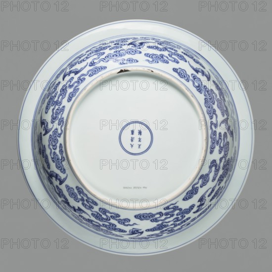Platter on stand, blue-and-white porcelain, Qing dynasty, Yongzheng period 1723–1735. The platter on a low foot with a wide rim sits on a carved wooden stand. Decorated with plant tendrils, bats, and Buddhist emblems. The center of the mirror has a rosette filled with an archaic swastika ornament. On the outer wall are bats among clouds. Porcelain vessel, height 21 cm, diameter 45.5 cm; China., A round ceramic bowl viewed from above with a smooth white interior and exterior, decorated on the inner wall with a repeating cloud-like pattern in blue, a thin blue rim around the top edge, and a circular blue double-ring near the center; the visible colors are white, blue, pale beige on the exposed foot rim, and a gray background.