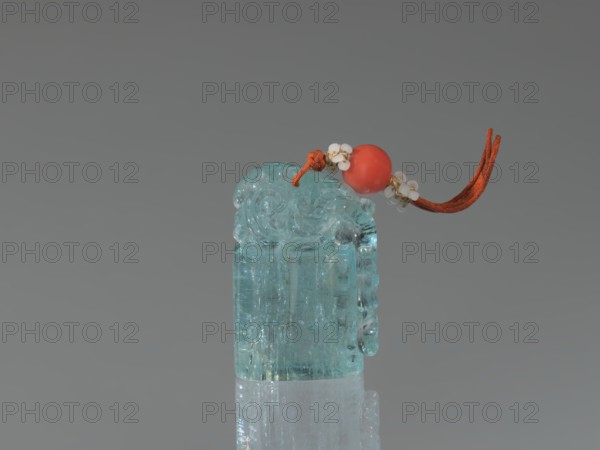 Seal stamp pendant, carved aquamarine stone, polished in the upper part; carved lion seal motif; strung with river pearls and coral; Qing dynasty 1644–1911, 18th–19th century; China; height 4.1 cm, width 1.8 cm, depth 2.7 cm., A translucent carved aqua-blue stone with rounded decorative carving on top sits upright on a reflective surface, topped by a short orange cord threaded through a smooth round coral-red bead and accented with small white bead clusters; the background is flat gray and the visible colors include aqua-blue, orange, coral-red, white, and gray.