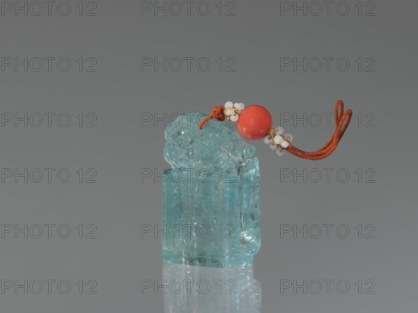 Seal stamp pendant, maker, Chinese; Qing dynasty 1644–1911; 18th–19th century. Carved aquamarine stone seal with a lion motif; upper part carved and polished. Strung with river pearls and coral. Dimensions: height 4.1 cm, width 1.8 cm, depth 2.7 cm., A translucent bluish-green carved object with a glossy surface sits upright and reflects below; on top of it rests a red-orange cord threaded through a smooth round red bead and accented with small white bead clusters, all set against a neutral gray background.