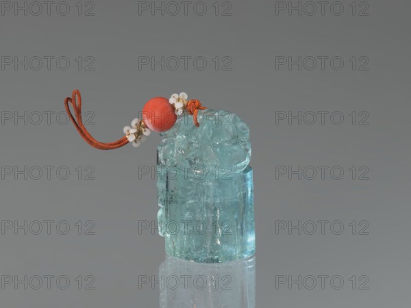 Seal stamp pendant, carved aquamarine stone, polished in the upper part; depicts a lion; strung with river pearls and coral. Chinese, Qing dynasty 1644–1911, 18th–19th century. Height 4.1 cm, width 1.8 cm, depth 2.7 cm., Seals; used as a seal stamp., A translucent blue-green carved object sits upright on a reflective surface, topped by a rounded decorative piece. Attached to the top is an orange cord looped and knotted, threaded through a smooth round bead in a bright coral-red color, and accented by a few small white round beads clustered with tiny gold-toned metallic spacers. The background is a uniform gray.
