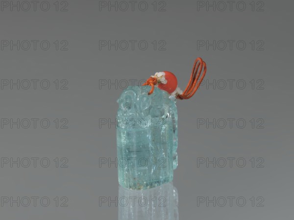Seal stamp pendant, carved aquamarine stone, carved and polished in the upper part; string with river pearls and coral. Qing dynasty 1644–1911, 18th–19th century, China. Subject: lion iconography; function: seal stamp. Dimensions: height 4.1 cm, width 1.8 cm, depth 2.7 cm., A translucent light blue carved stone pendant with rounded ornamental detailing at the top sits upright on a reflective surface, topped by a bright red bead and threaded with an orange cord loop and small white decorative accents; the background is a smooth gradient of gray.