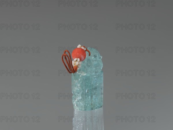 Seal stamp pendant, Chinese, Qing dynasty 1644–1911; carved stone aquamarine, polished in the upper part; carved lion iconography; drilled for suspension with string featuring river pearls and coral; 18th–19th century; seal stamp; dimensions: height 4.1 cm, width 1.8 cm, depth 2.7 cm., A small translucent carved object in a pale aqua color sits upright on a reflective surface, topped by a few decorative elements including a round bright coral-red bead, a few tiny white flower-shaped beads, and a looped cord in a rusty orange-brown hue; the background is a smooth gradient of gray.