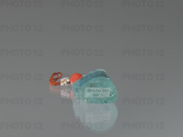 Seal stamp pendant, Chinese, Qing dynasty 1644–1911. Lion iconography. Stone aquamarine, carved and polished in the upper part; strung with river pearls and coral. Seal stamp, 18th–19th century. Dimensions: height 4.1 cm, width 1.8 cm, depth 2.7 cm., A small translucent teal object with a smooth, irregular shape rests on a reflective gray surface, accompanied by a looped orange cord, a round red bead, and a cluster of small white beads; the scene shows reflections of these items on the surface and the overall colors visible are teal, orange, red, white, and gray.