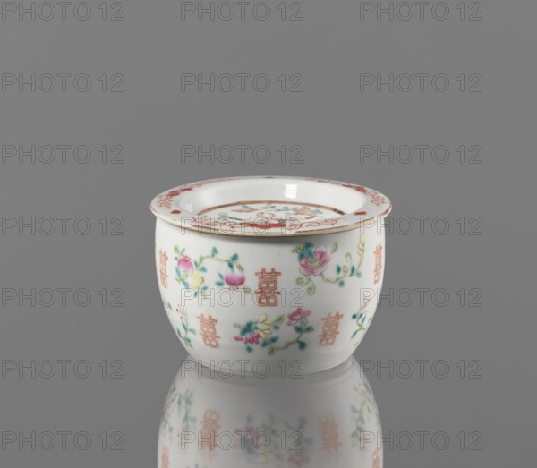 Hand warmer, ceramics, colored porcelain, Qing dynasty 1644–1911, Tongzhi period 1862–1874. Bulging vessel with a flat recessed lid pierced with five holes. Decorated with floral motifs — peonies, lotuses — peaches; longevity shou iconography. China, 1862–1874. Height 7.3 cm; diameter 11.2 cm., A small round porcelain bowl with a flared rim sits on a reflective surface against a plain gray background, showing a reflection beneath it. The bowl is primarily white with painted floral designs in pink, green, yellow, and turquoise, and decorative motifs in a reddish-orange around the rim and scattered on the body. The rim has a thin band of reddish-orange decoration and a narrow gold edge, and the interior of the bowl shows a circular painted scene with the same color palette. The painted flowers include blossoms and leaves with visible brushwork in shades of pink, green, yellow, and blue-green on the exterior. The overall scene is clean and evenly lit, emphasizing the glossy finish of the ...