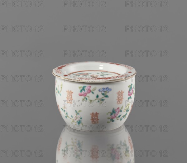 Hand warmer, purchase, Colored porcelain, Qing dynasty Tongzhi period, 1862–1874, China. Bulging vessel with a flat recessed lid pierced with five holes. Decorated with floral motifs — peonies, lotuses — and peaches as longevity shou iconography. Height 7.3 cm; diameter 11.2 cm., A small rounded ceramic cup with a flared rim sits on a reflective surface against a gray background, decorated with painted floral sprigs and repeating geometric motifs; the cup is primarily white with visible colors including pink, green, blue, yellow, and red, and the rim shows a red decorative border with a slightly worn edge.