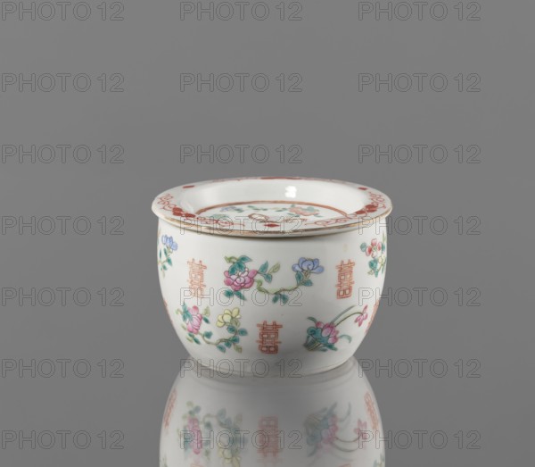 Hand warmer, colored porcelain, Qing dynasty Tongzhi period, 1862–1874, China. Ceramic vessel with bulging body and flat recessed lid pierced with five holes; decorated with floral motifs—peonies, lotuses—and peaches as longevity shou iconography. Height 7.3 cm; diameter 11.2 cm., A small white ceramic bowl with a flared rim sits on a reflective gray surface, showing a soft mirrored reflection beneath it; the bowl is decorated with delicate floral motifs in pink, blue, green, and yellow, accented with fine reddish-orange decorative marks along the rim and repeated vertical ornamental symbols around the body, and there are touches of gold along the rim.