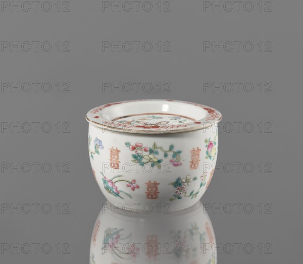 Hand warmer, Colored porcelain, Qing dynasty Tongzhi, 1862–1874. Bulging vessel with a flat recessed lid with five holes. Decorated with floral motifs—peonies, lotuses—and peaches; longevity shou iconography. China; height 7.3 cm, diameter 11.2 cm., A small white ceramic bowl with a flared rim sits on a reflective gray surface against a gray background; the bowl is decorated with delicate floral motifs in pink, green, blue, and yellow and has repeating geometric vertical motifs in orange around its exterior, while the rim features a red decorative band and a thin gold edge, and the bowl and its colorful decorations are reflected faintly on the surface below.