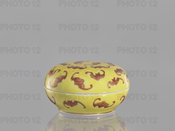 Round box with a convex lid a cinnabar vessel for seals, bats iconography, ceramics, colored porcelain, Qing dynasty, Tongzhi 1862–1874, Chinese, lid, seals, vessel; height 4 cm, diameter 7 cm., A small rounded box with a lid sits on a reflective surface; the box is yellow with a repeating decorative motif in reddish-brown, and the background is a uniform light gray.